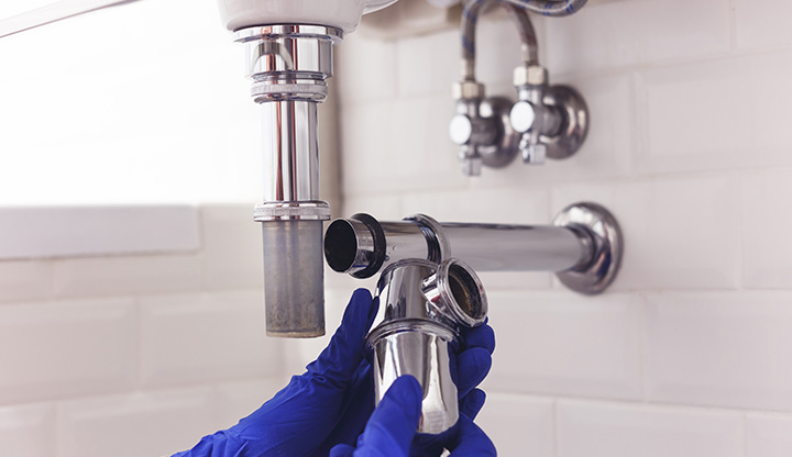 Hands wearing blue gloves holding a chrome plumbing trap beneath a sink 설치